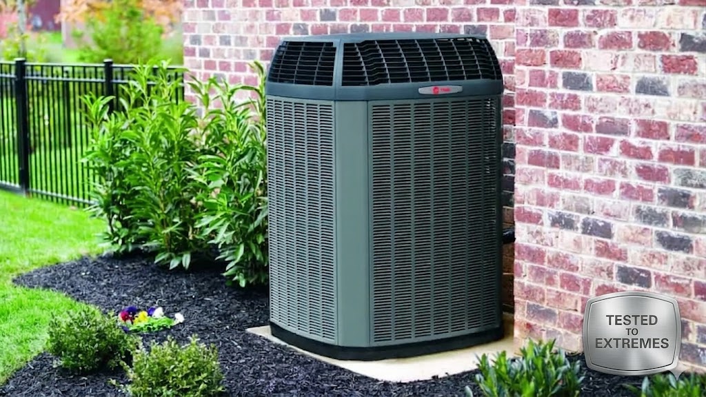 A large outdoor air conditioning unit is installed next to a brick house, surrounded by mulch, plants, and grass. Learn more about its reliability with the "TESTED TO EXTREMES" badge in the corner.